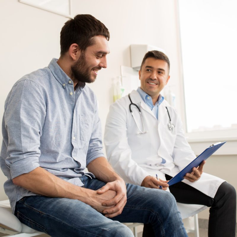 medicine-healthcare-people-concept-smiling-doctor-with-clipboard-young-man-patient-meeting-hospital
