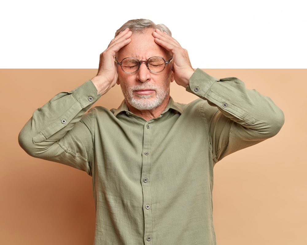 Displeased grey haired man suffers headache keeps hands on head to reveal pain needs painkillers has migraine after noisy party wears formal shirt isolated over brown background. Stressed pesnioner
